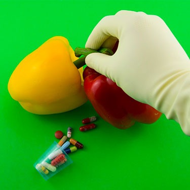 Una mano con guanto che sistema due peperoni, uno giallo e uno rosso, ai piedi dei due ortaggi c'è un bicchierino di pillole che si spargono lungo lo sfondo verde a tinta unita. 